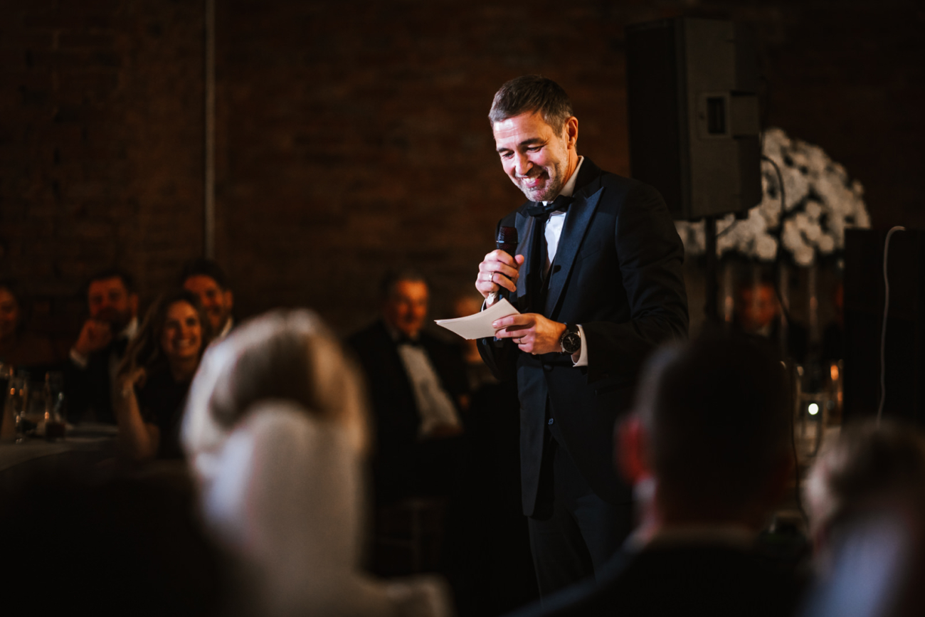 A best man in a tuxedo smiles as he delivers his wedding speech, microphone in one hand and his notes in the other, while guests laugh at the reception.