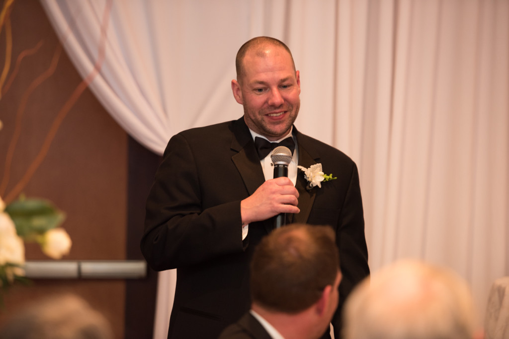 A groomsman in a tuxedo holds a microphone and smiles mid-speech at a wedding reception.